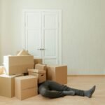 A person lying under a pile of moving boxes in a minimalistic room conveys tiredness from packing.